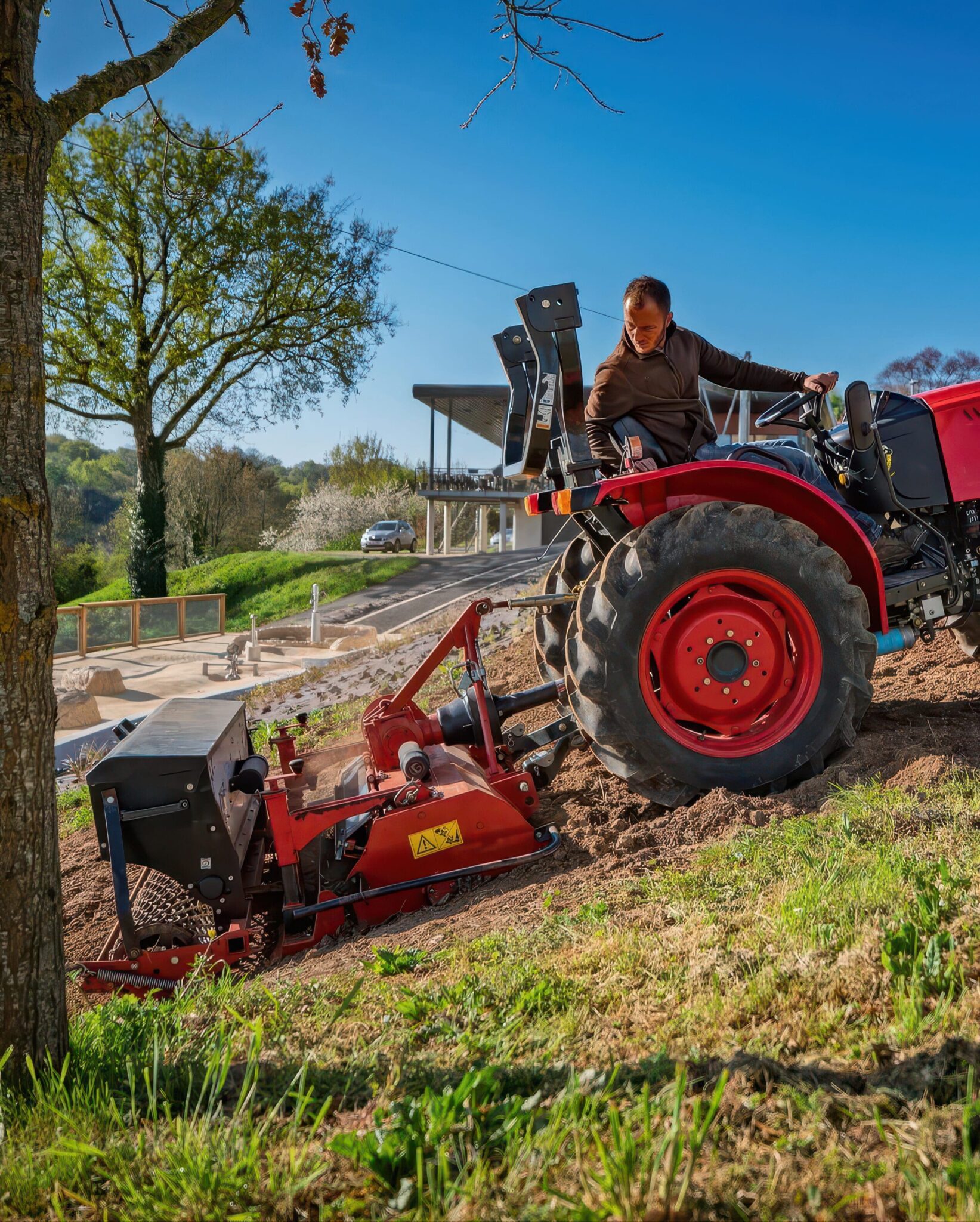 Paysagiste conduisant un tracteur rouge équipé d'un outil de travail du sol en pente