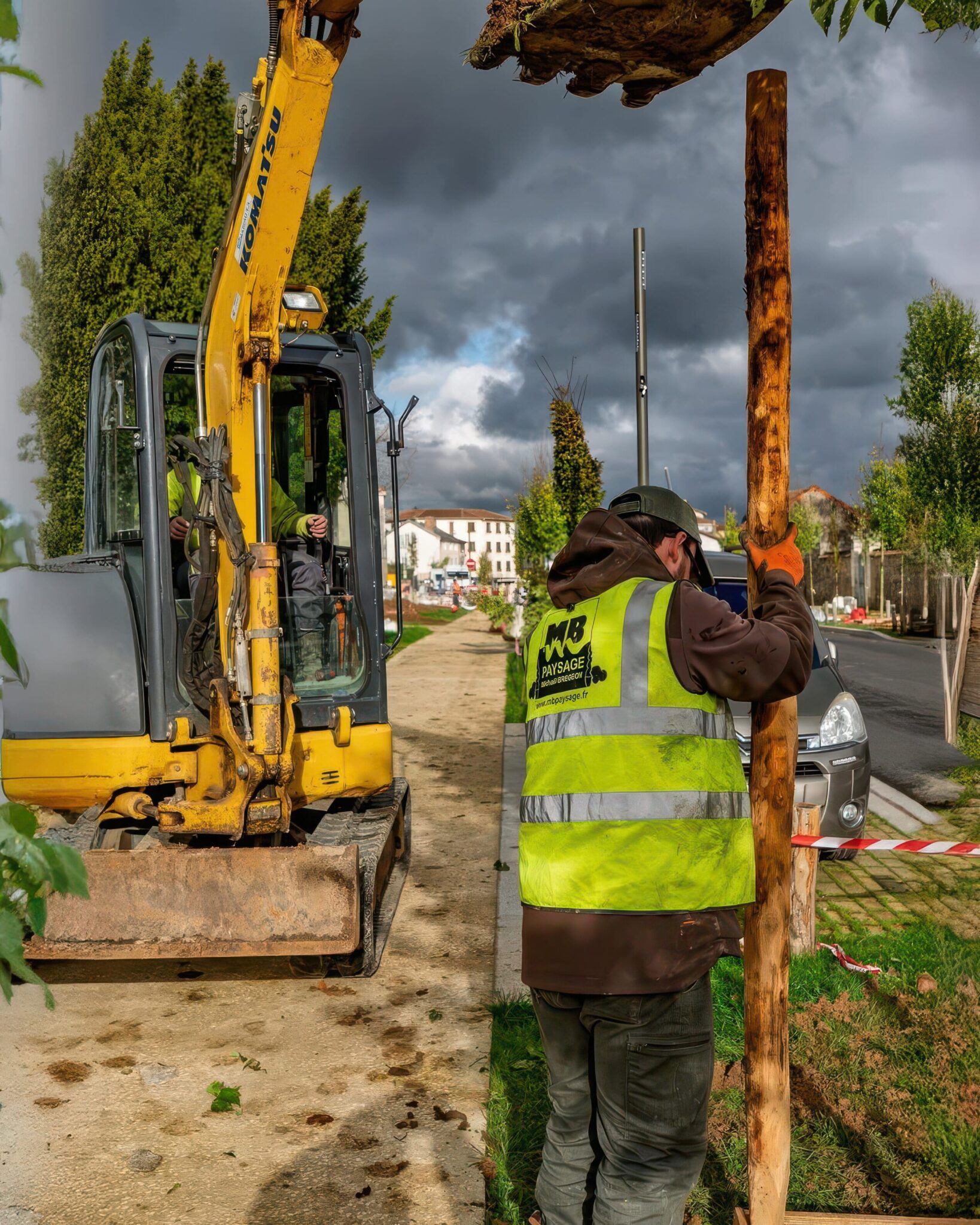 Paysagiste en gilet jaune posant un poteau en bois avec une pelleteuse jaune à côté