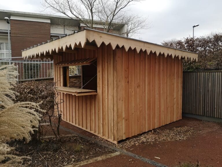 Cabane en bois clair avec un grand auvent décoré, dans un jardin
