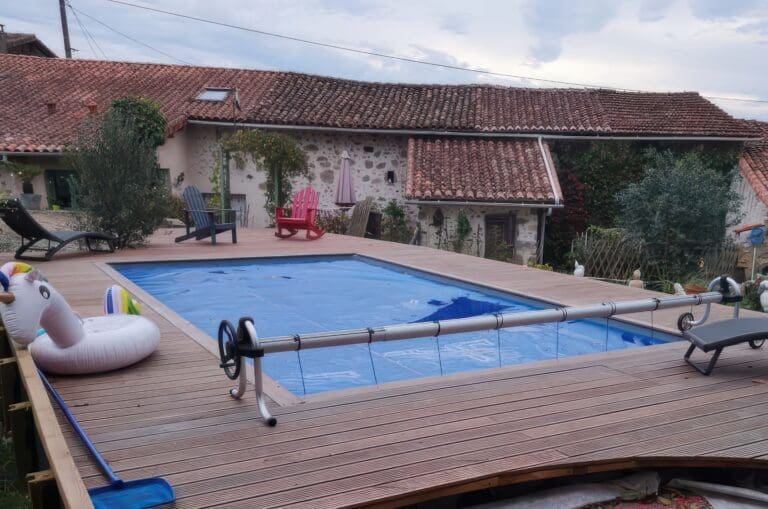 Piscine rectangulaire avec couverture bleue entourée d'une terrasse en bois avec un bouée licorne, chaises et transat