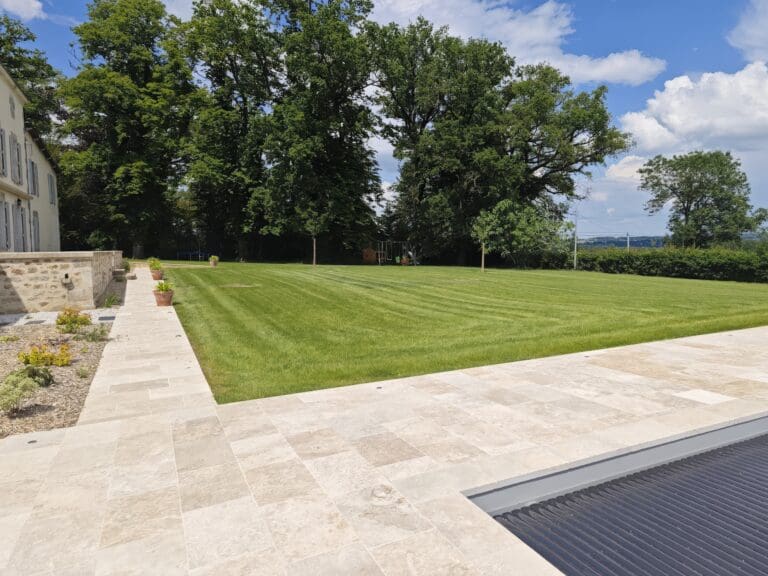 Grand jardin pelouse verte bordée de dallage clair avec maison en pierre à gauche
