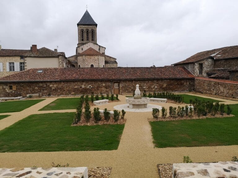 Jardin aménagé avec pelouses, allées gravillonnées, haies basses et fontaine sur fond d'anciennes bâtisses en pierre