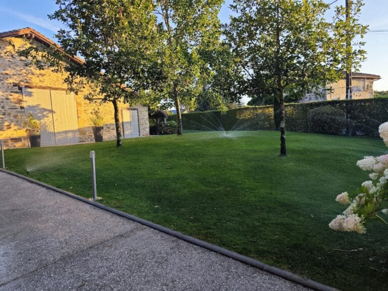 Pelouse verte avec arrosage automatique et arbres devant une maison en pierre au coucher du soleil