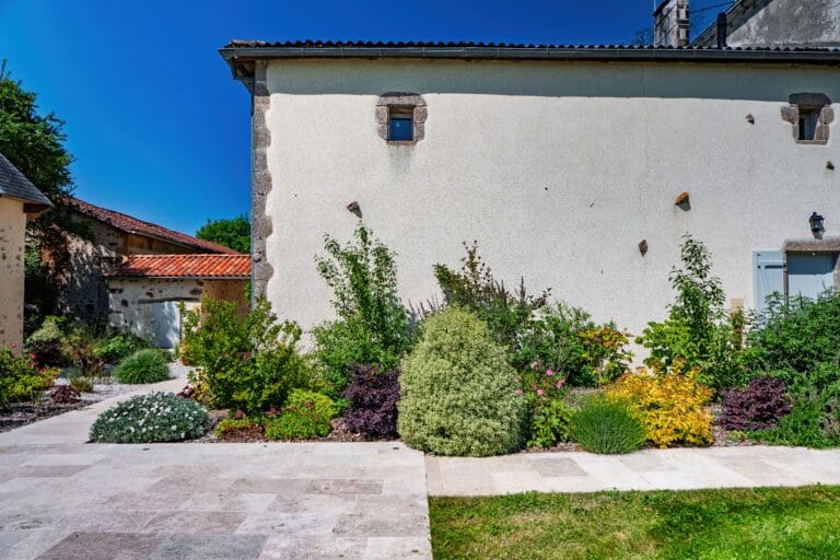 Parterre de buissons et arbustes devant un mur extérieur de maison en pierre sous un ciel bleu