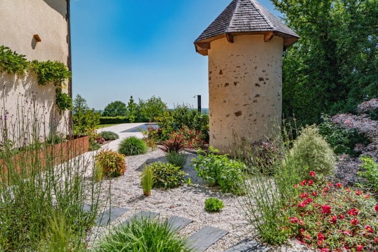 Jardin aménagé avec gravier blanc, plantes variées et petit bâtiment rond au toit en ardoise