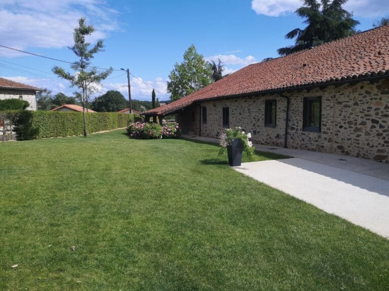 Jardin bien entretenu avec pelouse, haie, arbre et bordure fleurie devant une maison en pierre