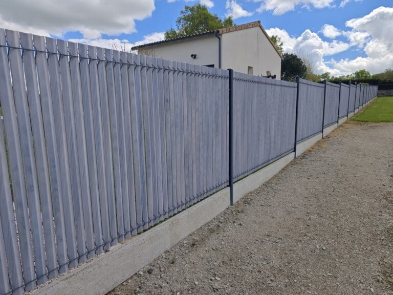 Clôture moderne en bois peint gris avec base béton devant une maison sous un ciel partiellement nuageux