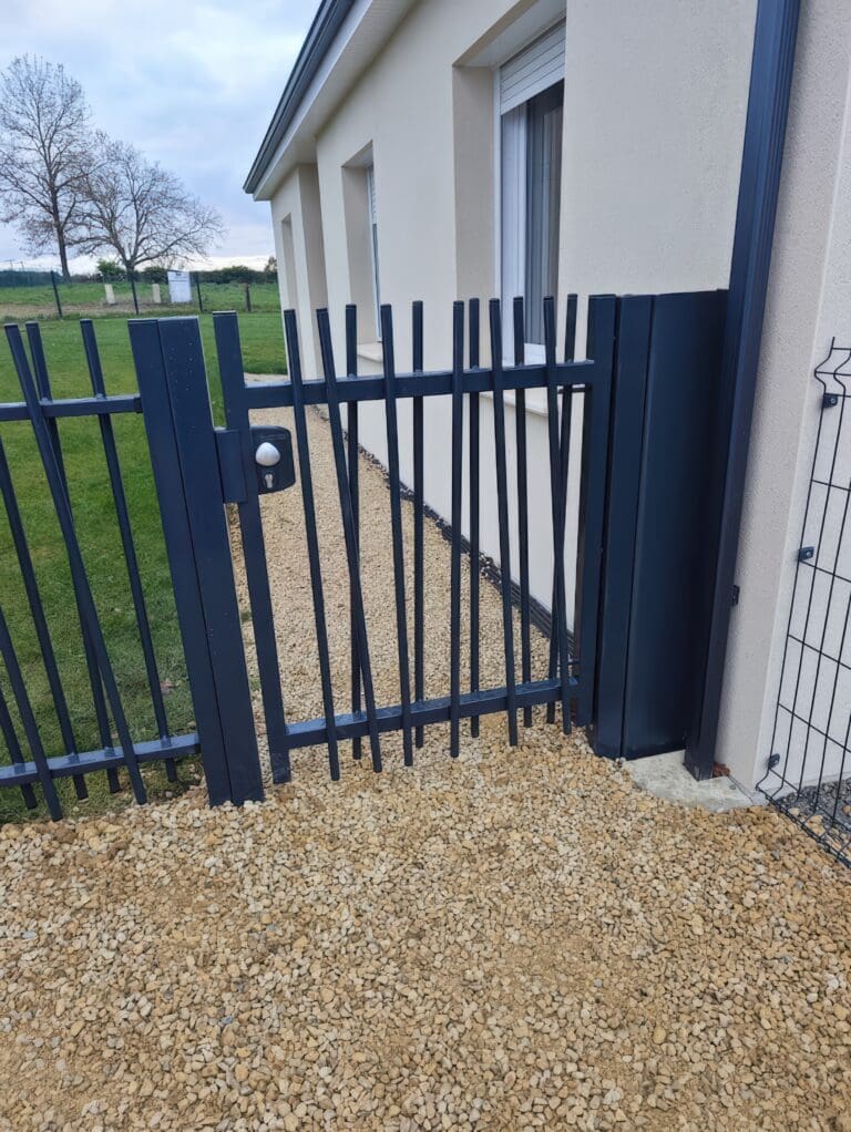 Portillon métallique noir avec des montants irréguliers devant une maison avec allée gravillonnée