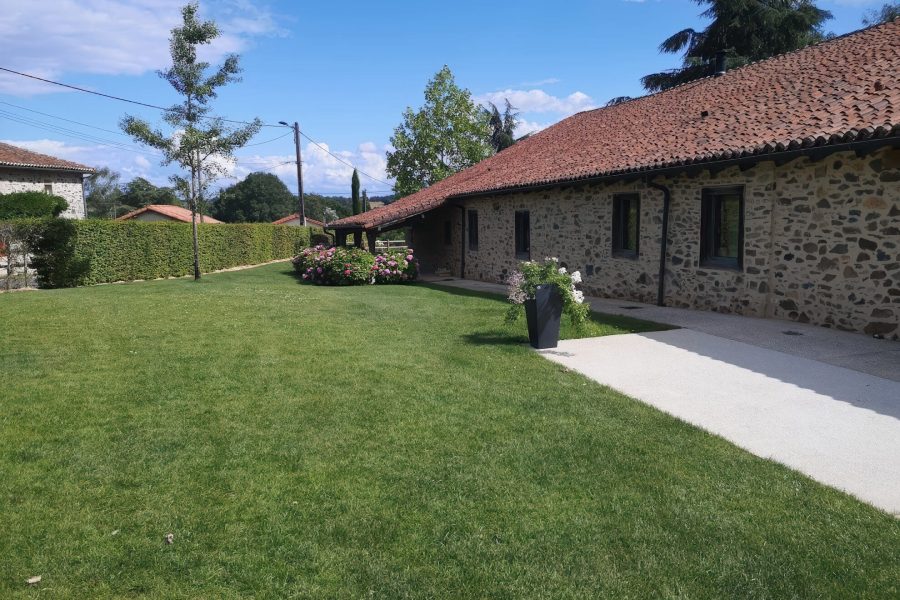 Jardin bien entretenu avec pelouse, haie, arbre et bordure fleurie devant une maison en pierre