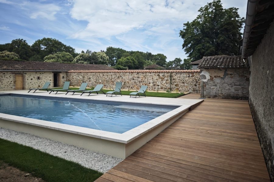 Piscine entourée d'une terrasse en bois avec six chaises longues et un mur en pierres en arrière-plan